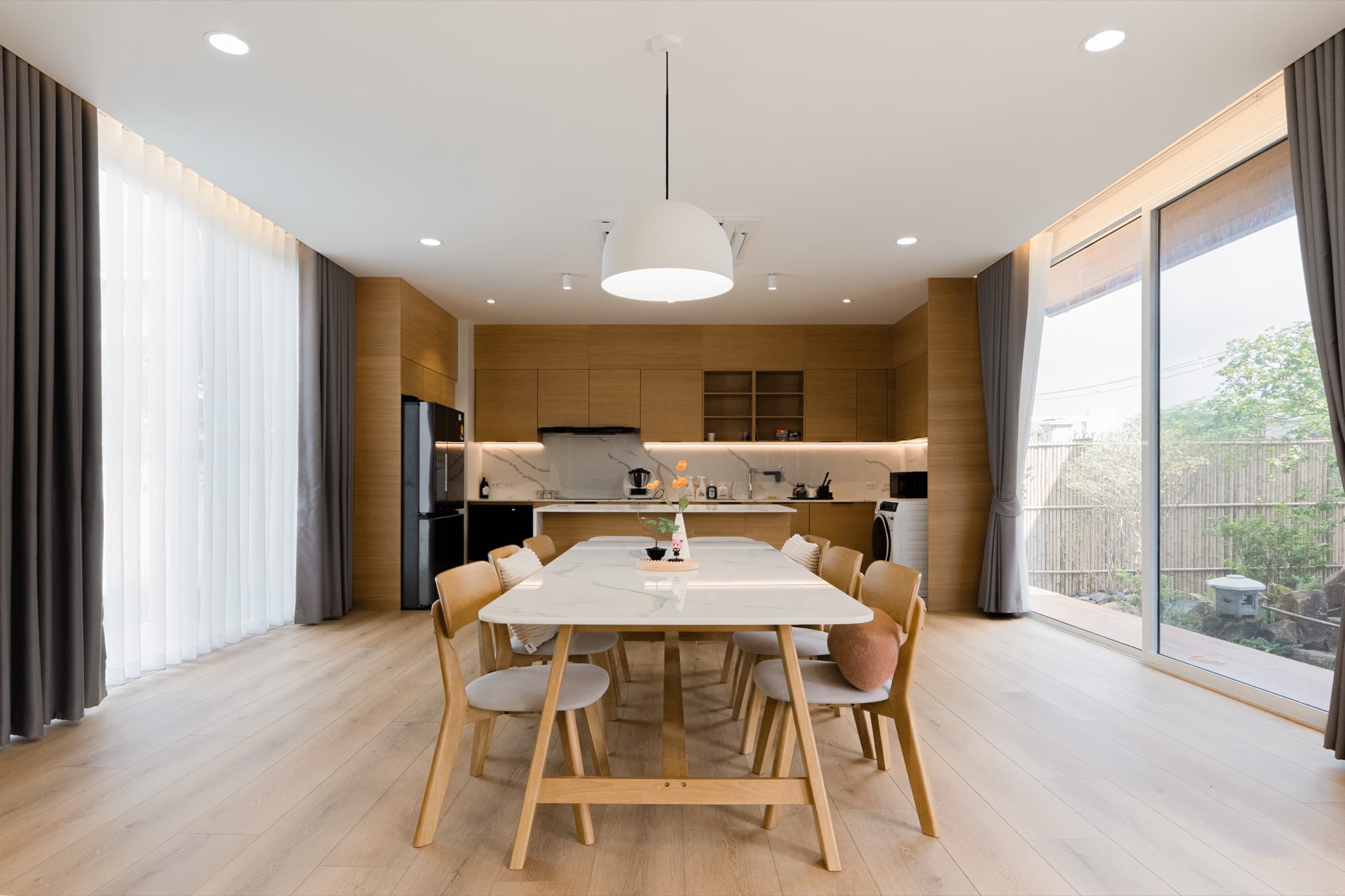 มุมโต๊ะทานข้าวไม้ยาวพร้อมเก้าอี้ 6 ตัว โคมไฟห้อยทรงกลม เชื่อมกับครัวไม้ด้านหลังและหน้าต่างบานใหญ่ ผลงานออกแบบภายในโดย OBJ Design & Build