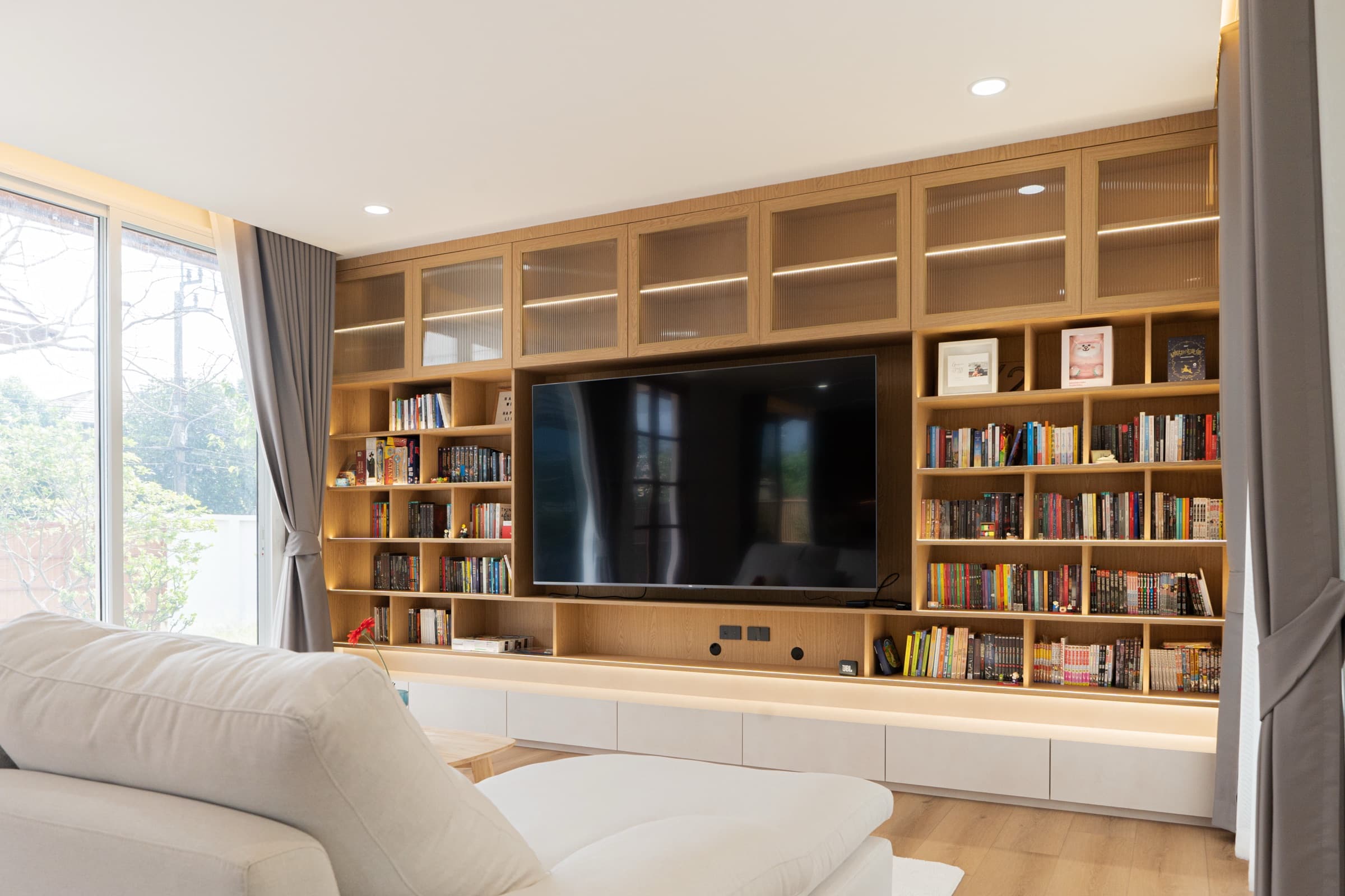 ห้องนั่งเล่นผนังทีวีไม้บิ้วท์อินเต็มผนัง มีชั้นหนังสือโดยรอบทีวี โซฟาผ้าสีขาวด้านหน้า ผลงานออกแบบภายในโดย OBJ Design & Build