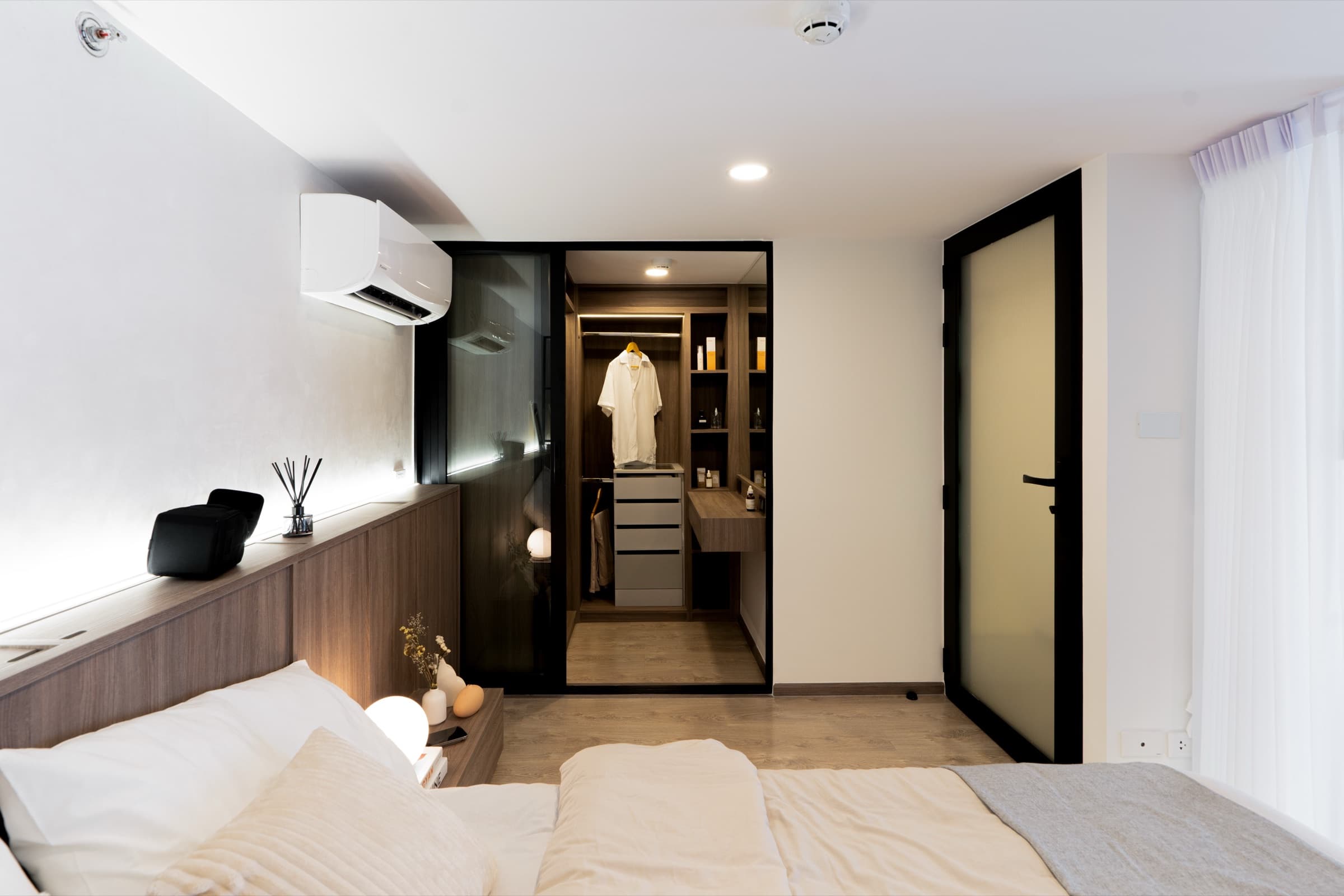 ห้องนอนอีกมุม เตียงพร้อมแอร์ติดผนัง มองเห็นตู้เสื้อผ้า walk-in ผ่านประตูกระจกกรอบดำ เสื้อแขวนพร้อมใช้งาน ผลงานออกแบบภายในโดย OBJ Design & Build